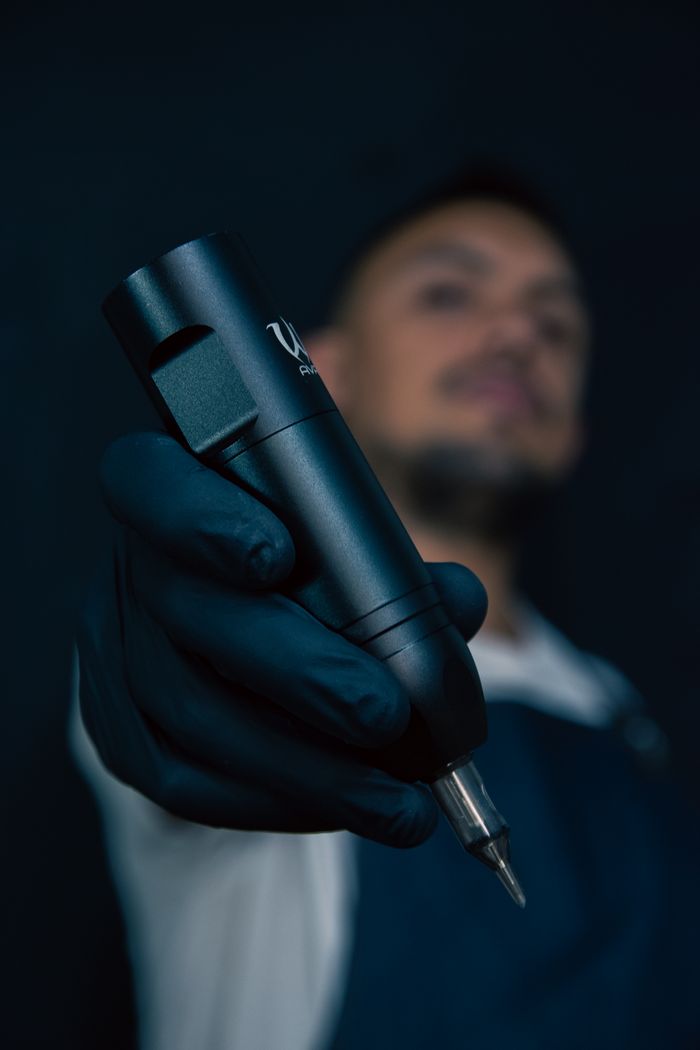 about-02 Tattoo artist wearing gloves holds a sleek tattoo machine in focus with blurred face.