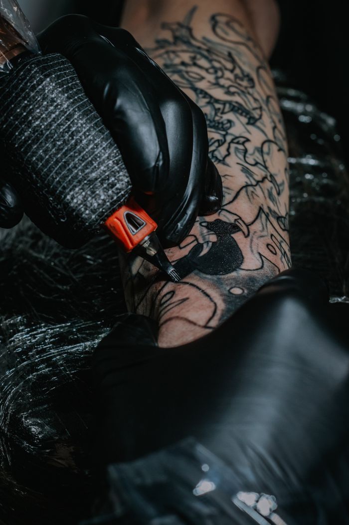 our-experience Intricate black tattoo being inked by a gloved artist, showcasing detailed artistry on arm.