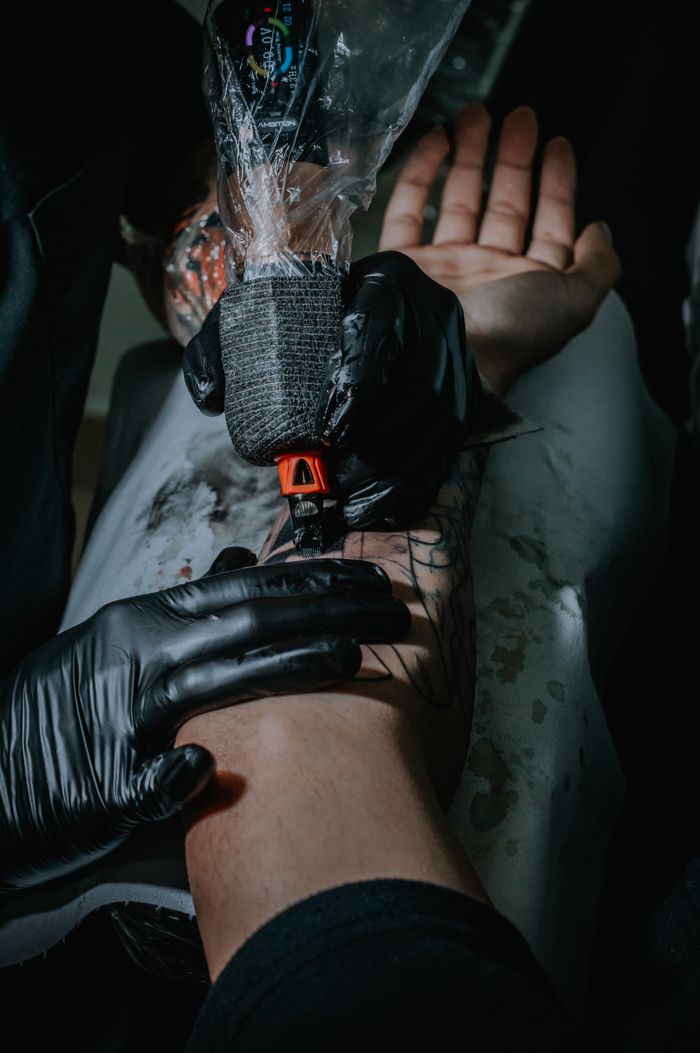 services-01 Tattoo artist working meticulously on a client's arm tattoo. Close-up of the tattoo process with gloves and tattoo machine.
